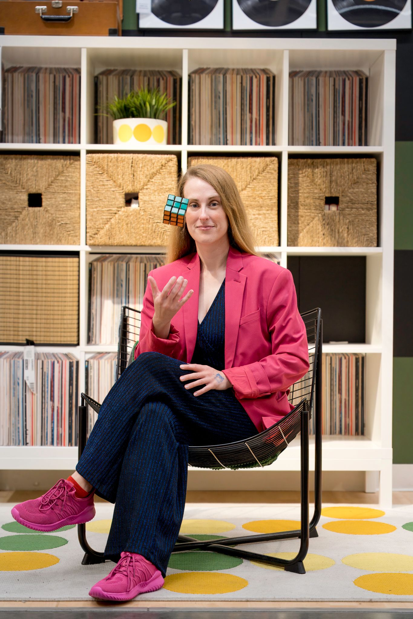 Woman in pink blazer and wide-leg jeans sits in chair holding Rubik's cube before shelving unit with vinyl records.