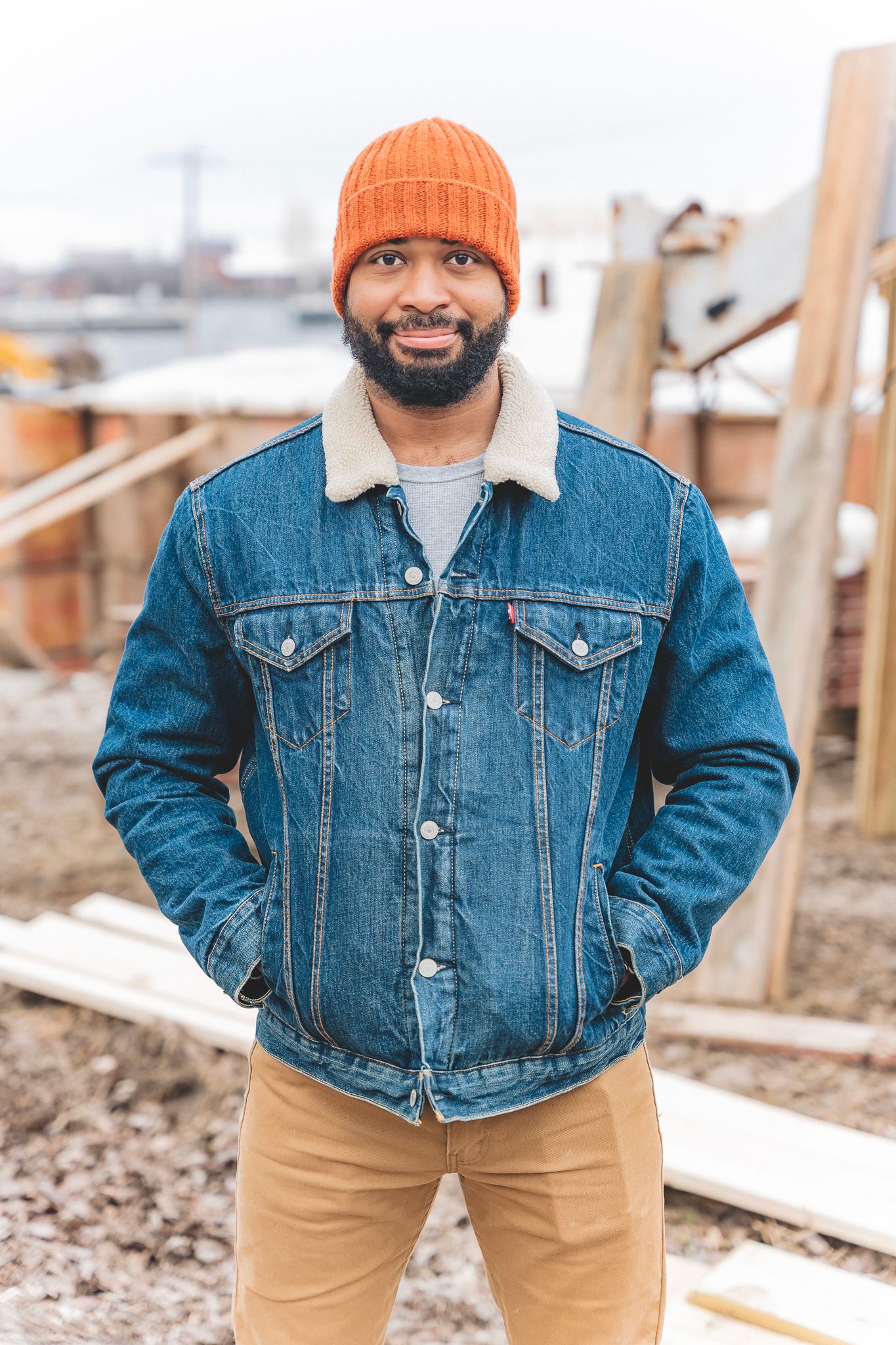 Smiling bearded man wearing an orange beanie and Levi denim jacket stands at an outdoor construction site.