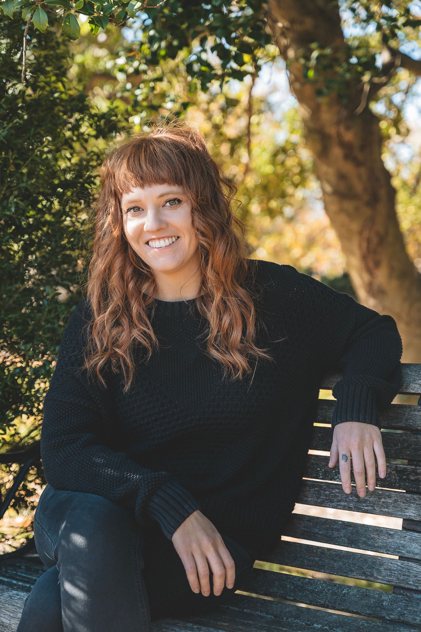 Smiling young woman with red hair sitting on a bench outdoors in a black sweater.