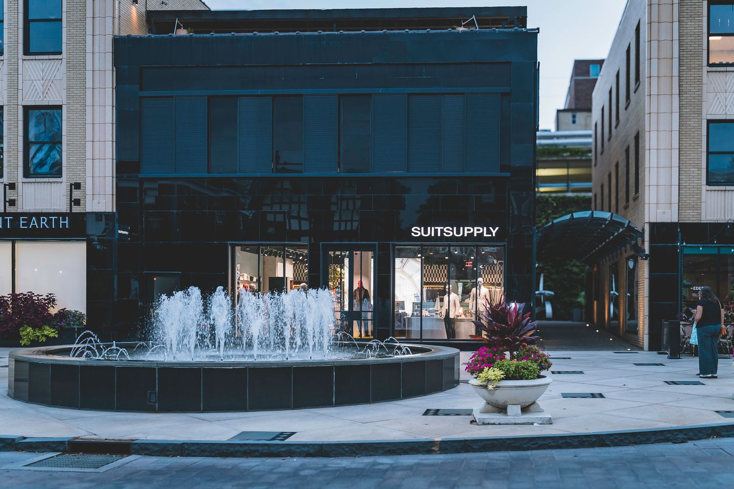 Urban plaza fountain outside Suitsupply store with modern architecture at dusk.