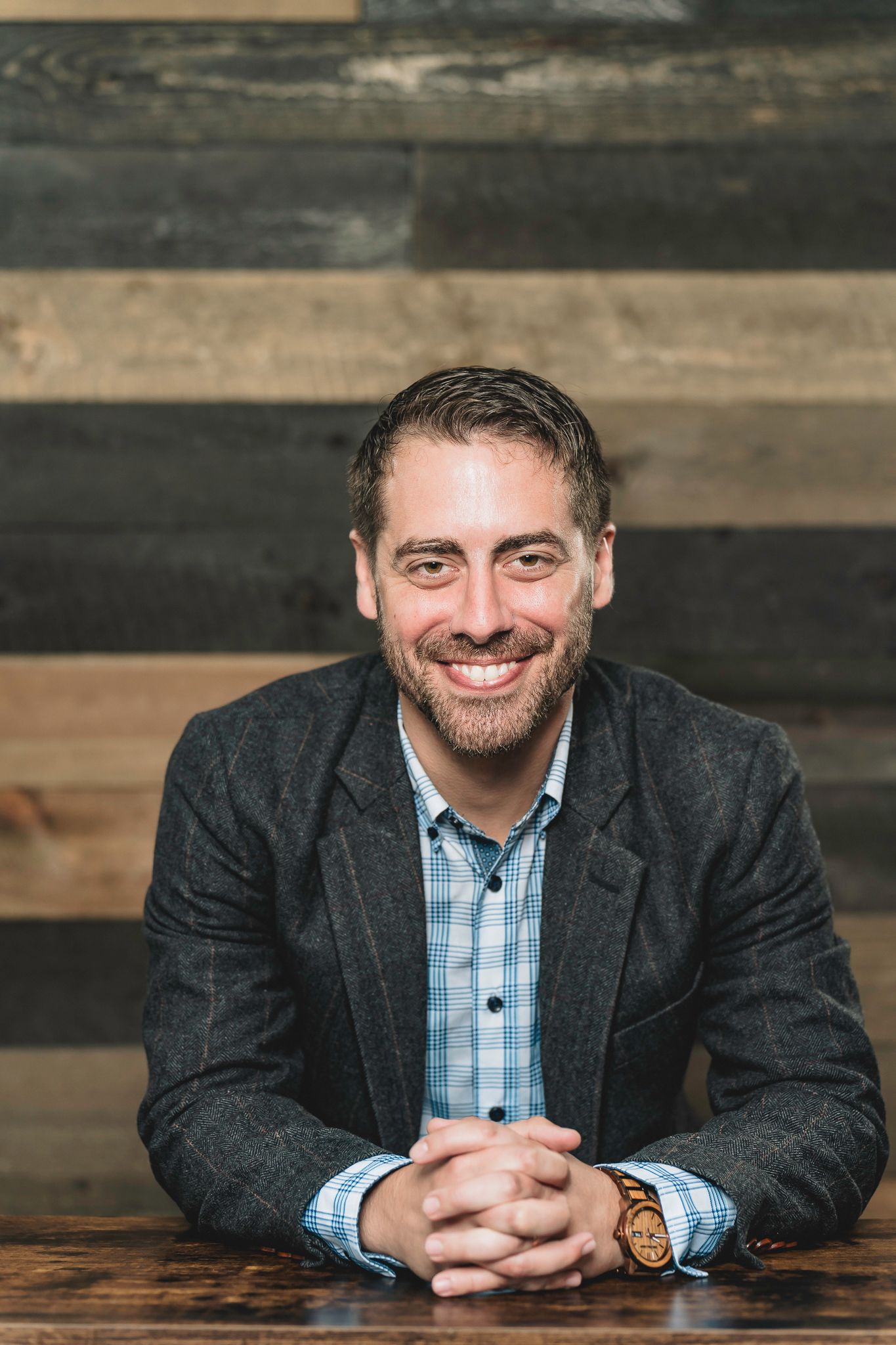 Smiling bearded man in gray blazer and blue shirt seated at wooden table with rustic wood plank wall background.