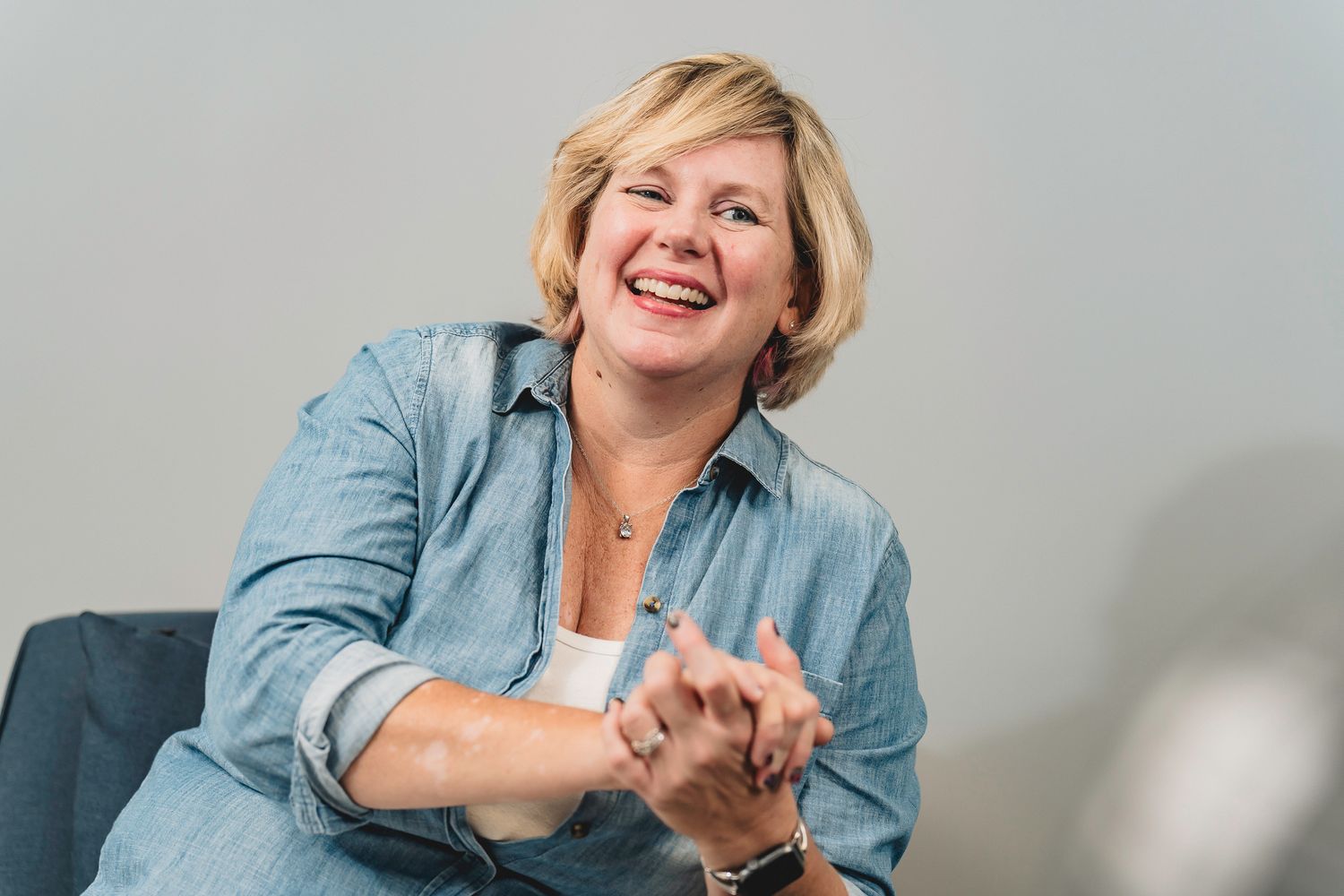 Cheerful blonde woman in denim shirt laughs warmly while seated in a casual professional setting.