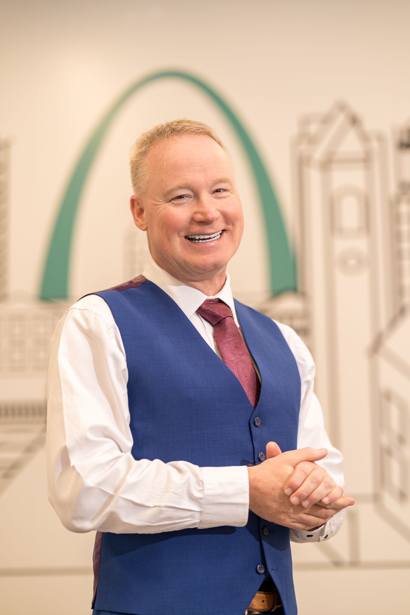 Smiling man in blue vest and pink tie posing before St. Louis Gateway Arch mural.