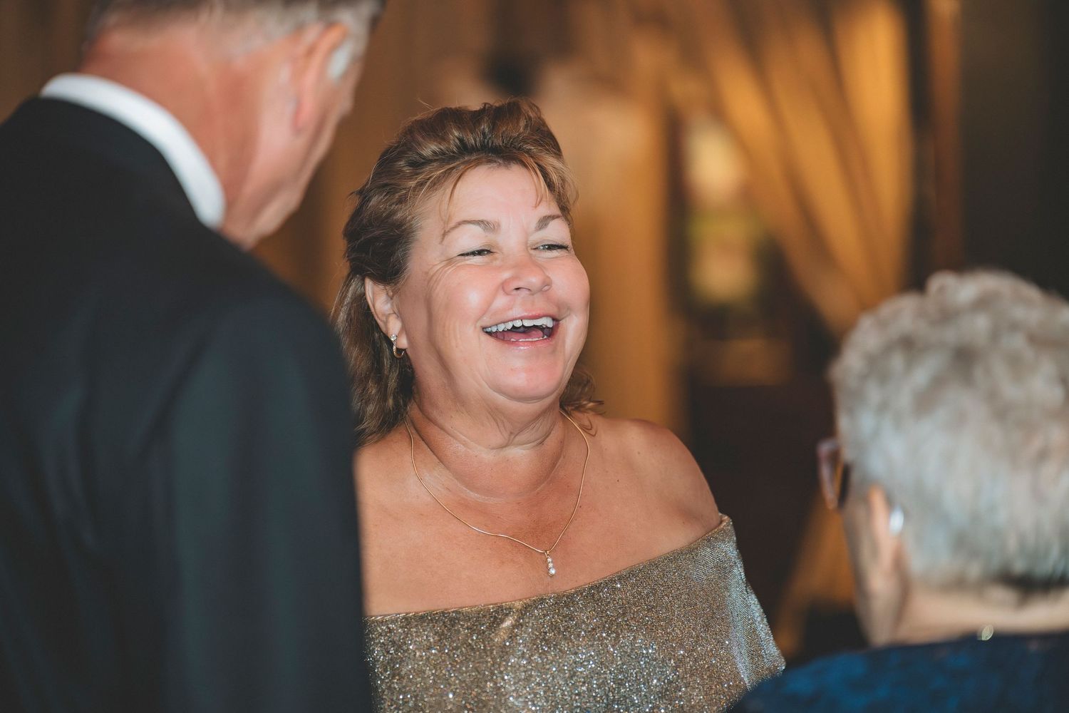 A joyful woman in a gold off-shoulder dress laughs warmly while conversing with guests at a wedding.