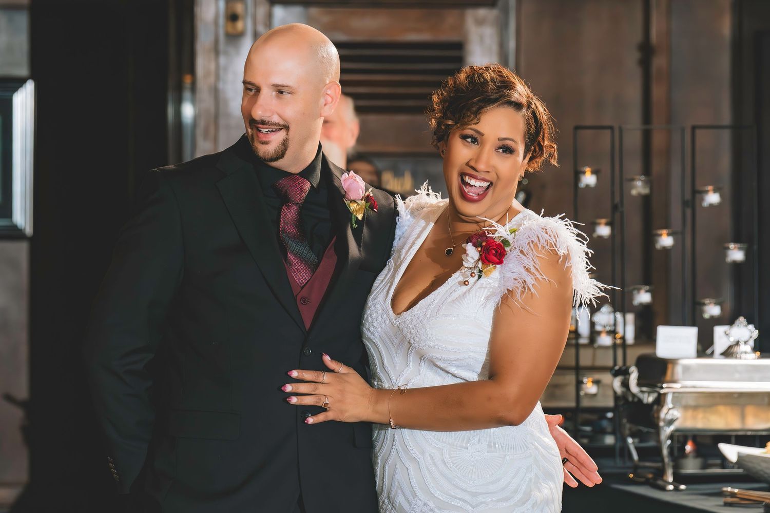 Smiling bride in white feathered gown and groom in dark suit pose together at wedding reception.