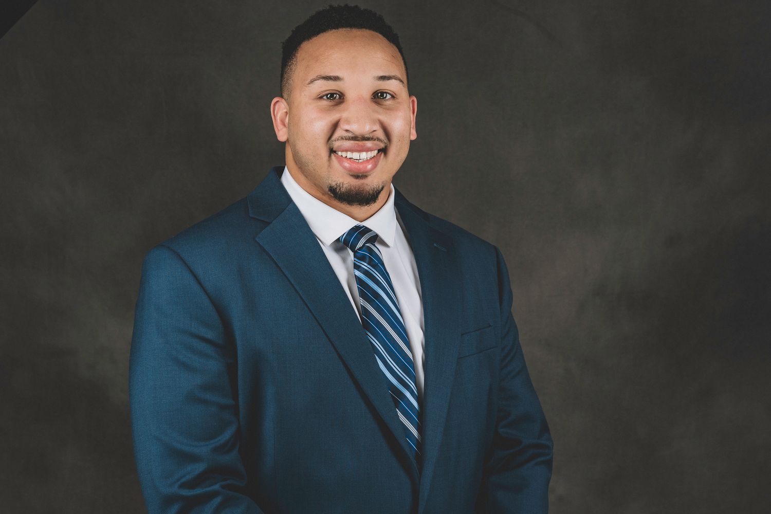 Smiling professional man in navy blue suit and striped tie posing against a dark gray studio background.