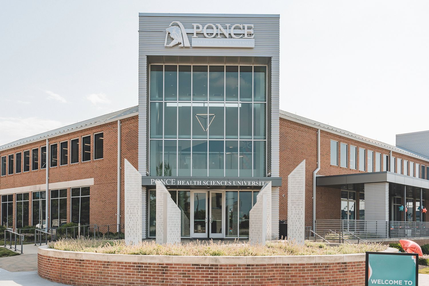 Modern brick and glass Ponce Health Sciences University building exterior with welcome sign on sunny day.