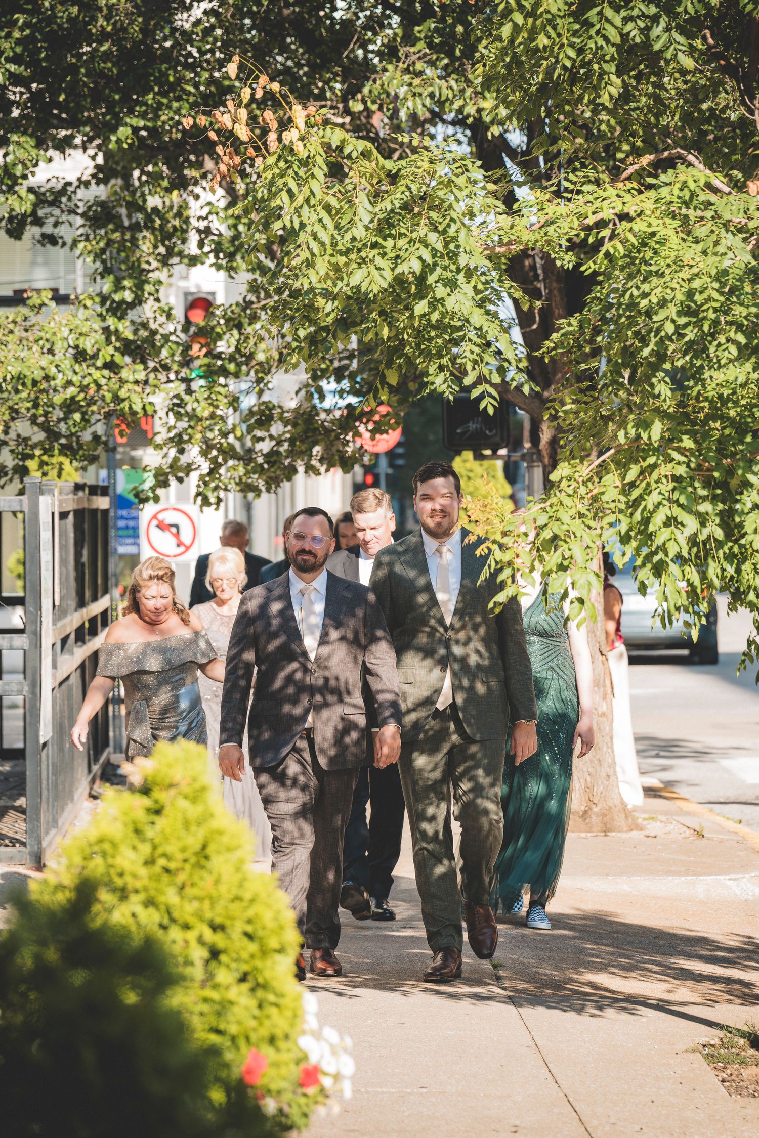 Wedding party guests in formal attire walk along a sunny tree-lined sidewalk toward a venue.