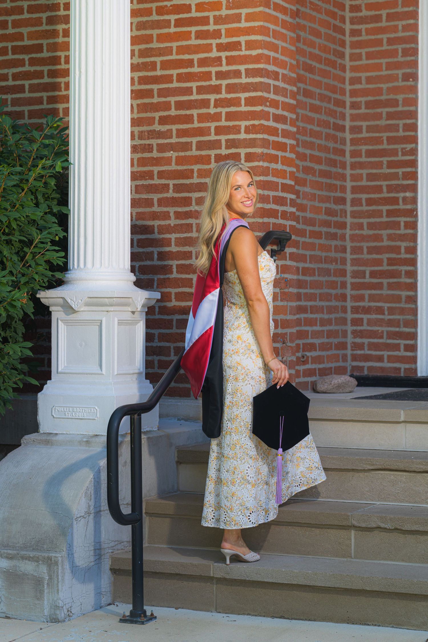 Blonde woman in floral dress carries graduation cap on brick building steps.