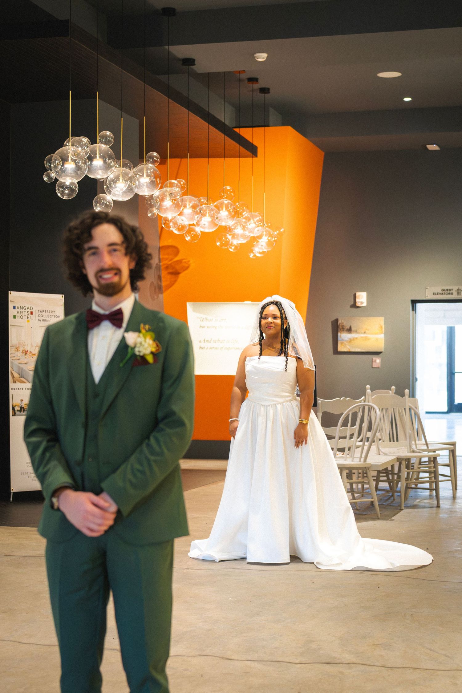 Groom in green suit stands as bride in white wedding dress approaches from behind.