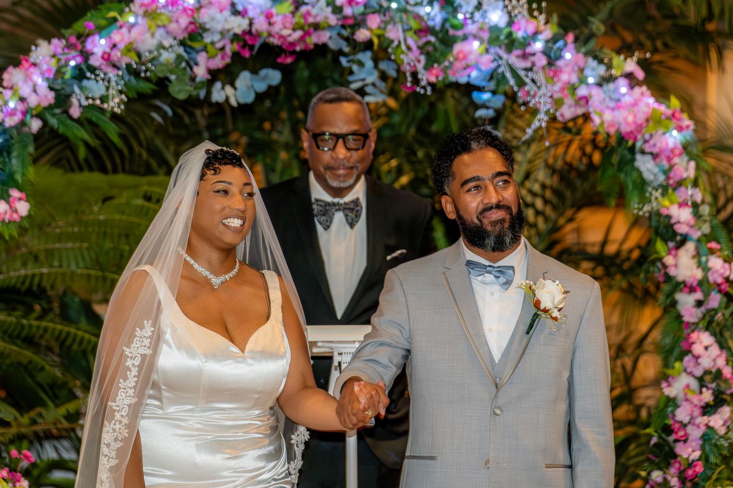 Smiling newlyweds hold hands with officiant in tuxedo under purple floral arch at night ceremony.