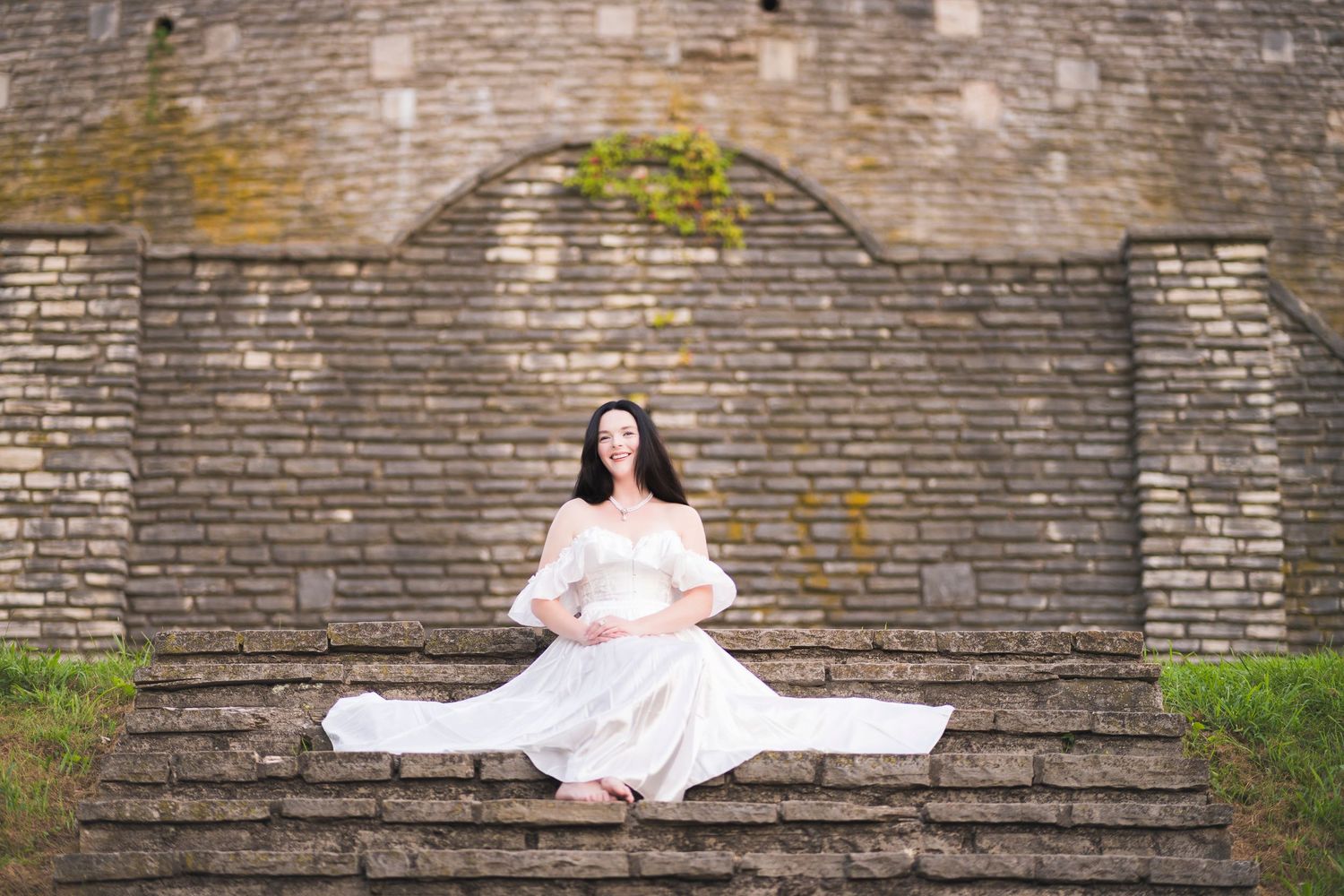 Woman in white off-shoulder dress sitting on stone steps in front of brick arch wall.