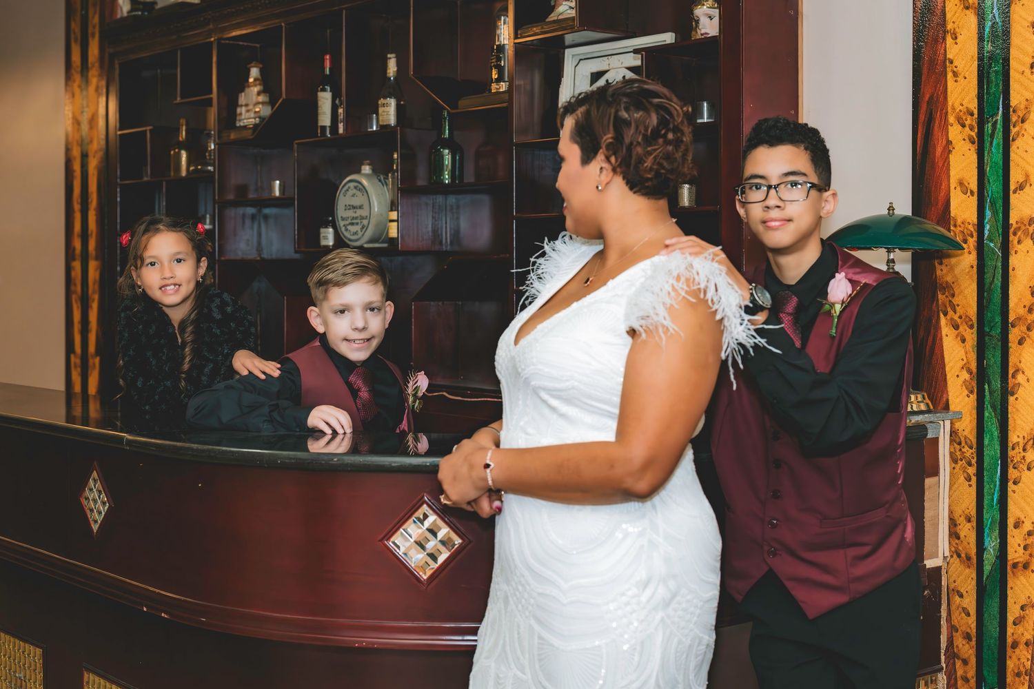 A bride in a white feathered dress stands with three children in burgundy and black formal wear.