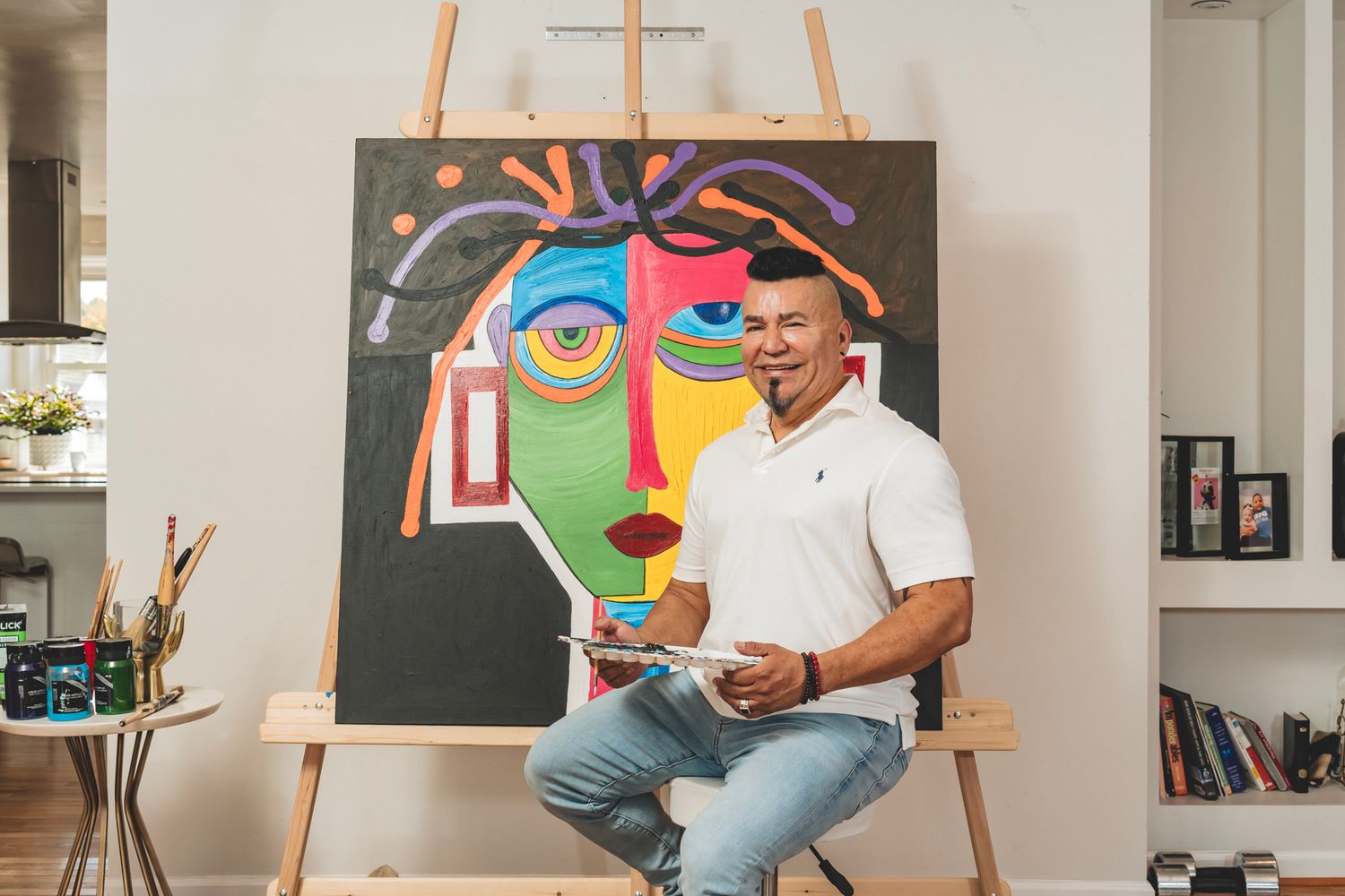 Artist smiling in front of colorful abstract face painting on easel in bright home studio.