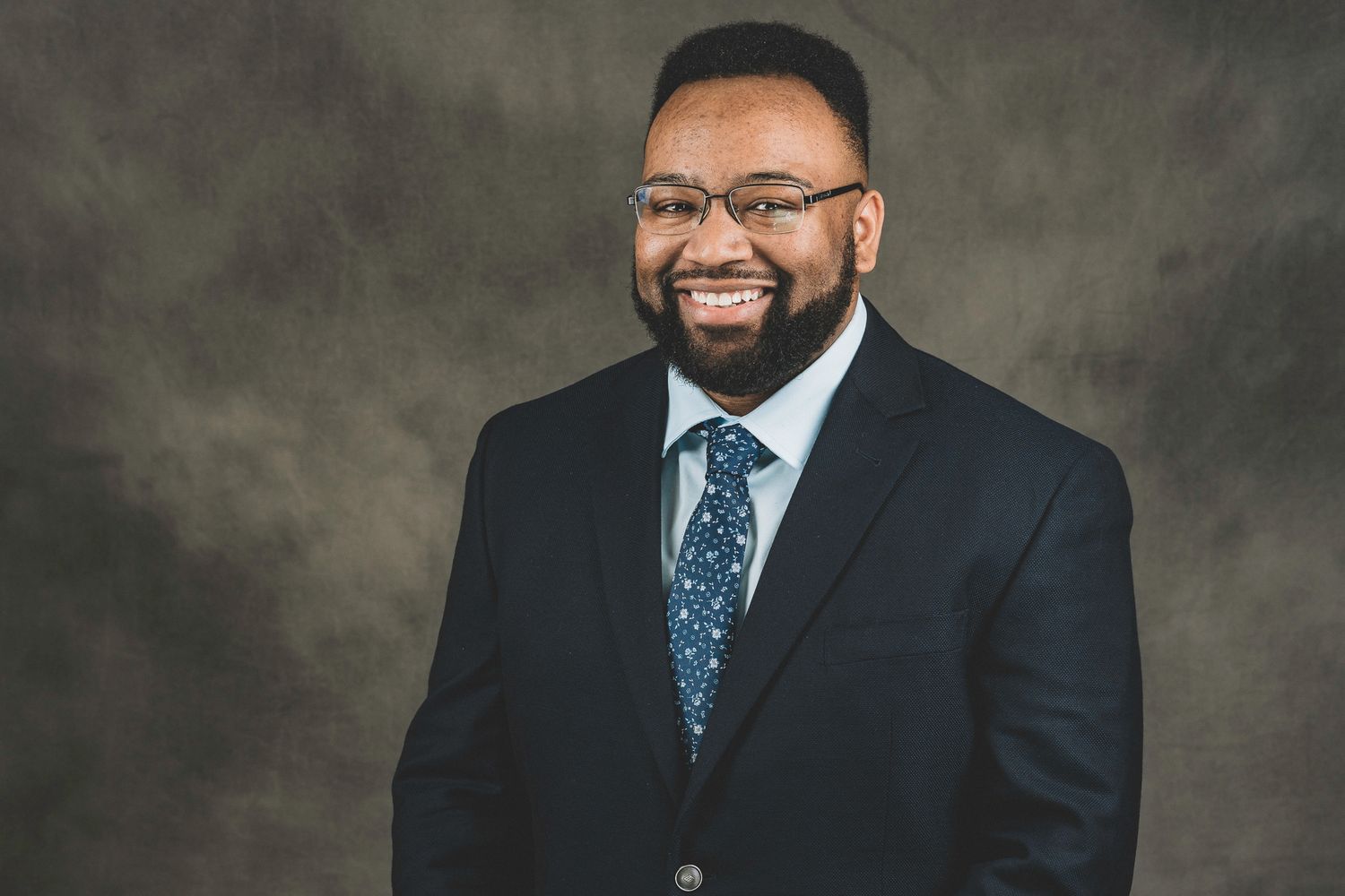 Smiling bearded man in black suit and patterned tie posing against a gray studio background.