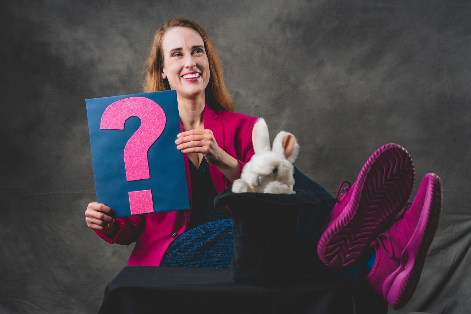 Woman in pink blazer holds question mark sign beside white rabbit in magician hat.