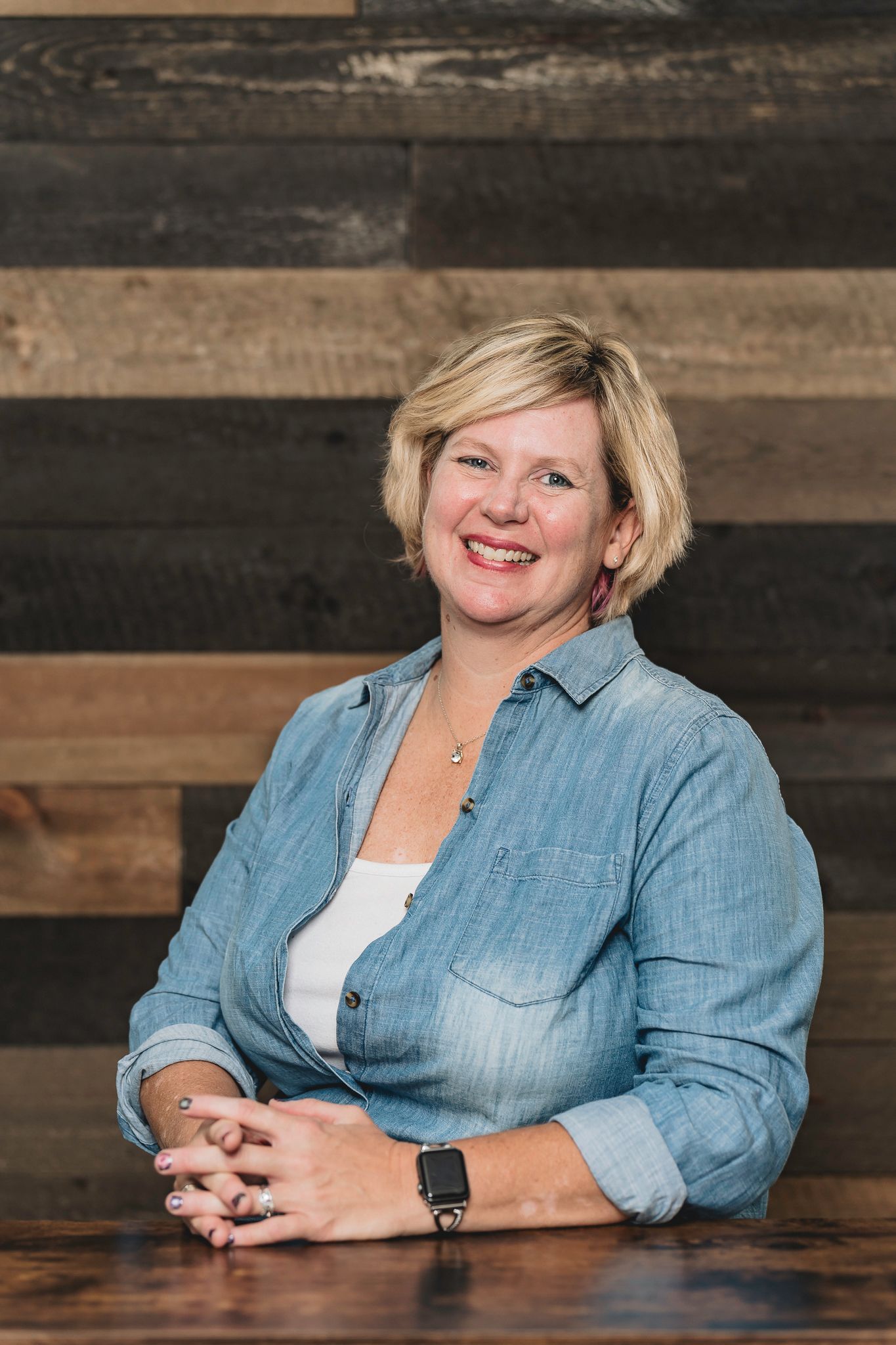 Smiling blonde woman in denim shirt seated at wooden table with rustic reclaimed wood plank wall background.