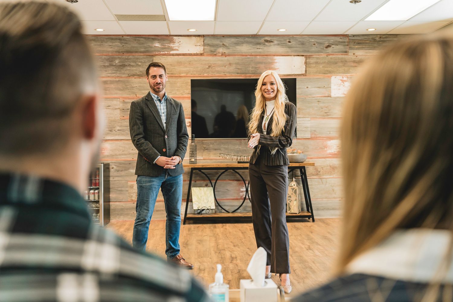 Man and blonde woman in business attire present to seated audience in rustic wood-paneled office space.