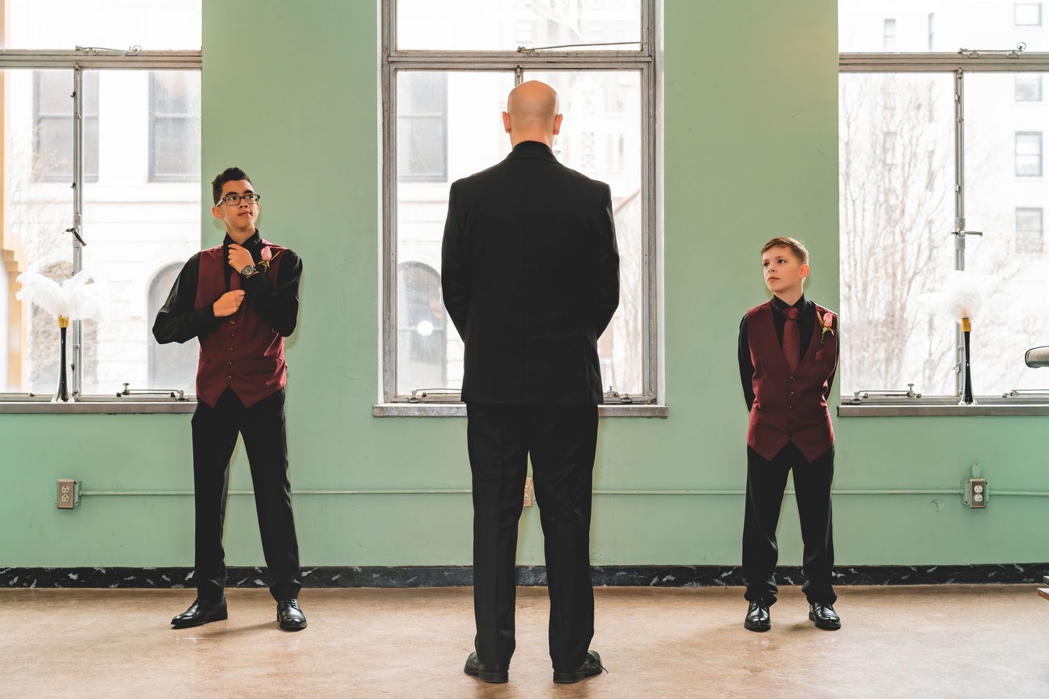 Groom stands between two groomsmen in burgundy vests in a bright windowed venue.