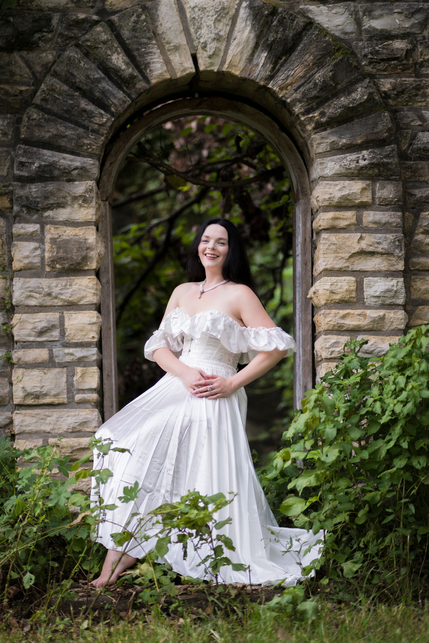Smiling woman in white off-shoulder dress posing in stone archway surrounded by green foliage.