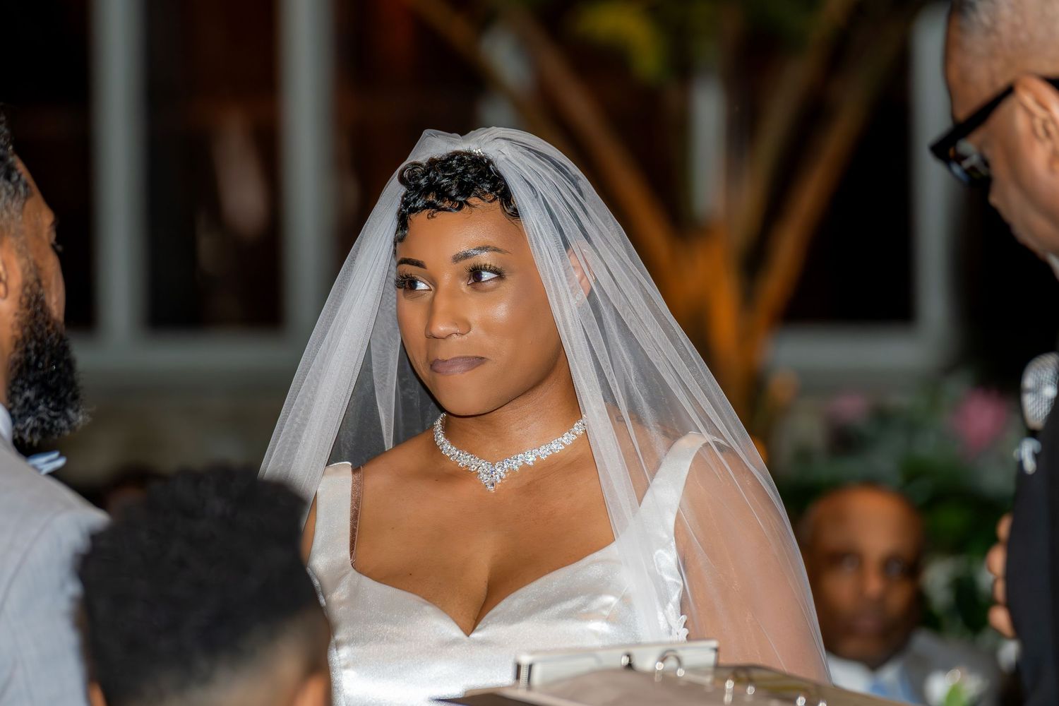 Elegant bride in white gown and veil wearing diamond necklace at outdoor wedding ceremony.