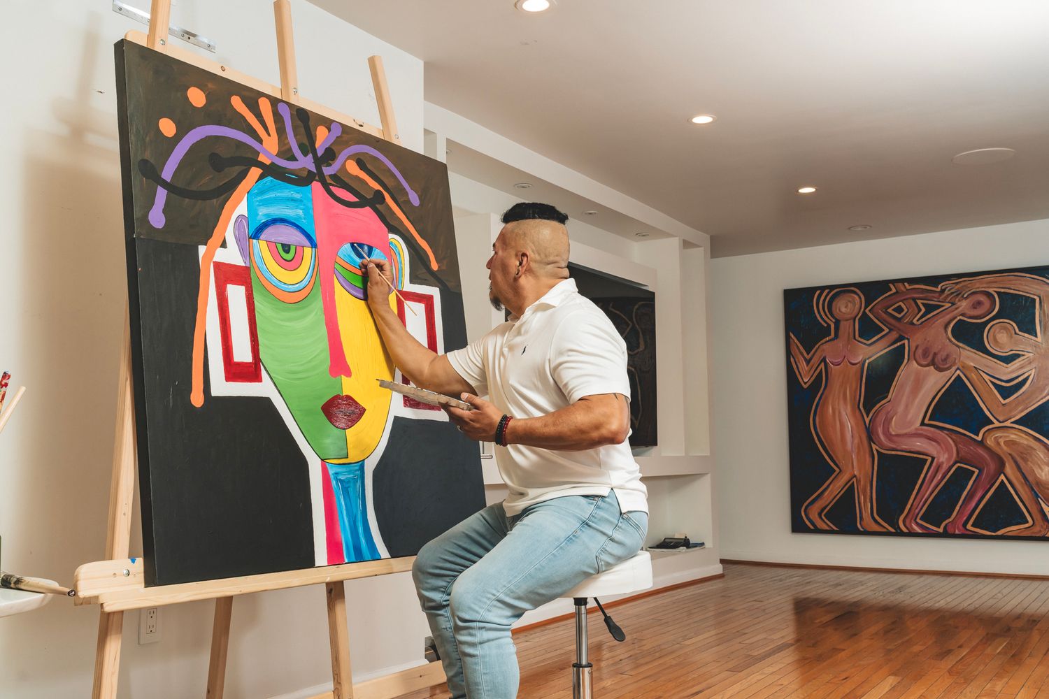 Male artist painting colorful abstract face on large canvas in bright art studio with gallery walls.