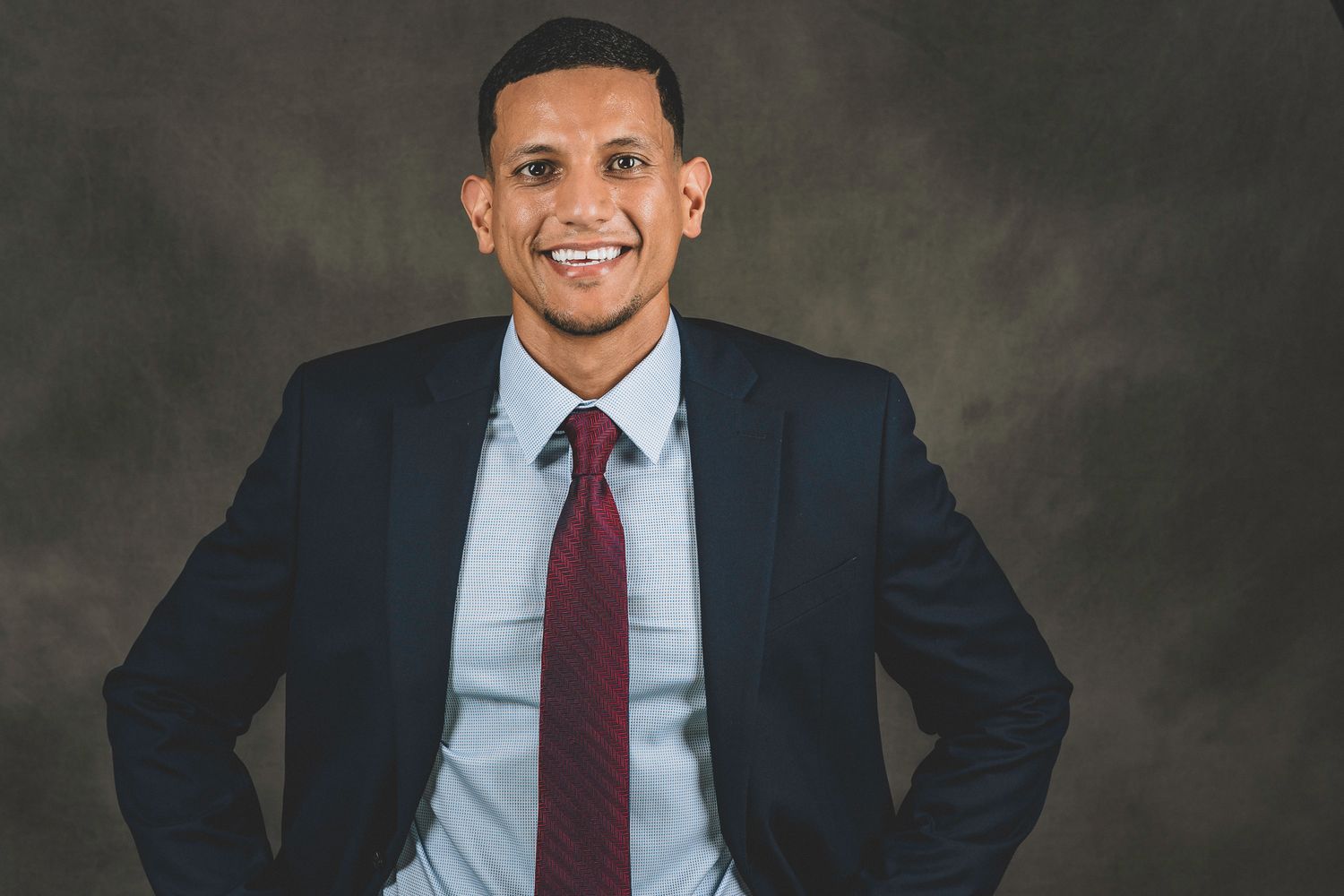 Smiling professional man in navy suit and red tie standing with hands on hips against gray studio background.