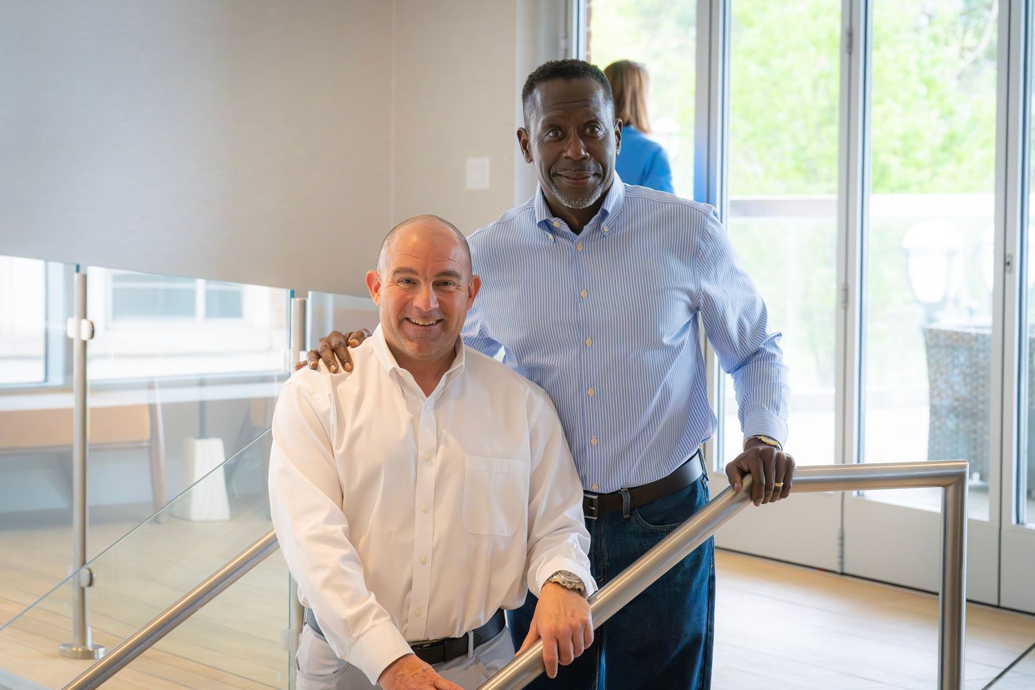 Two smiling businessmen posing together on a modern office staircase railing for a photo.