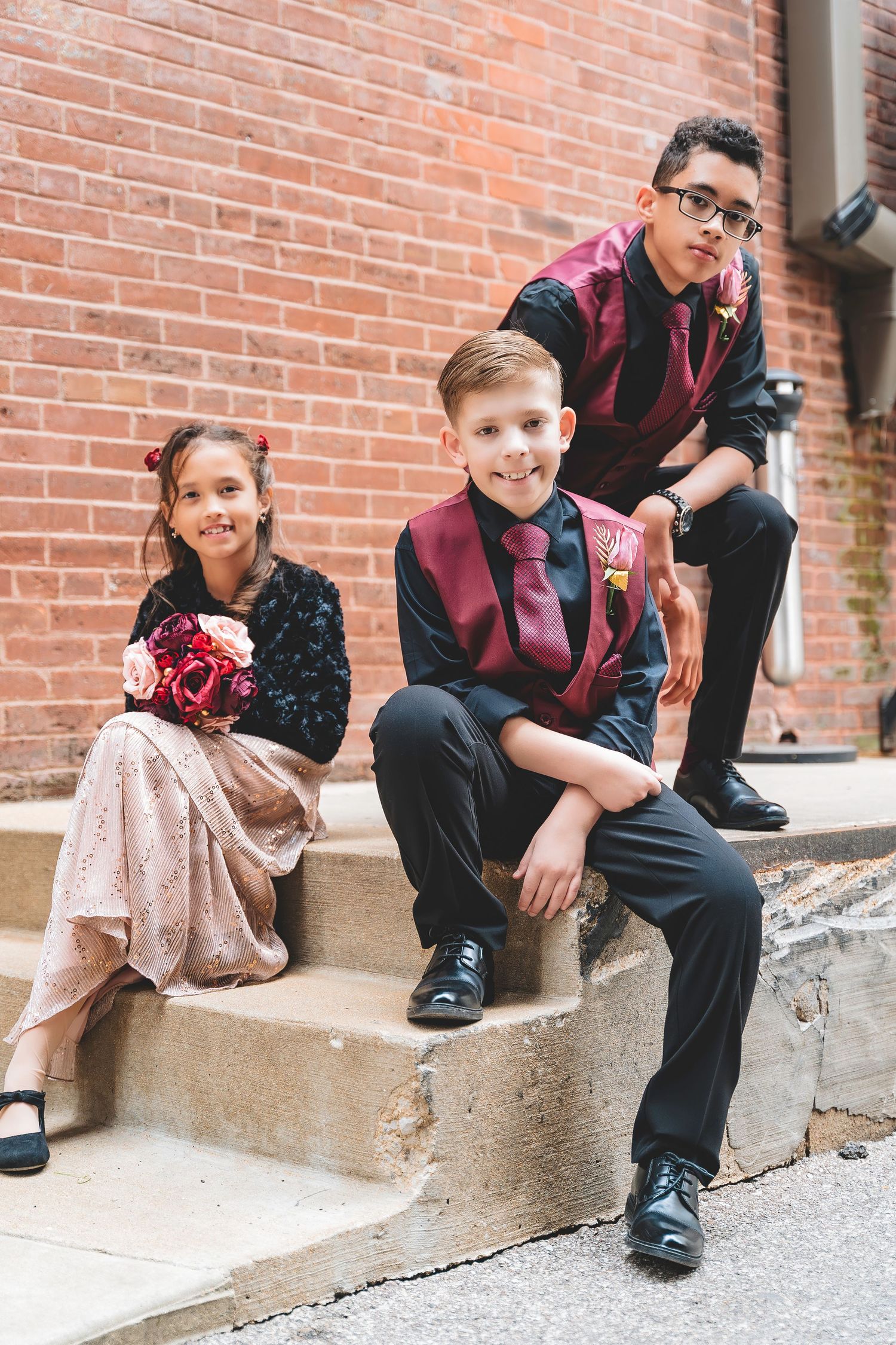Three children in formal burgundy and black wedding attire sit on outdoor brick steps.