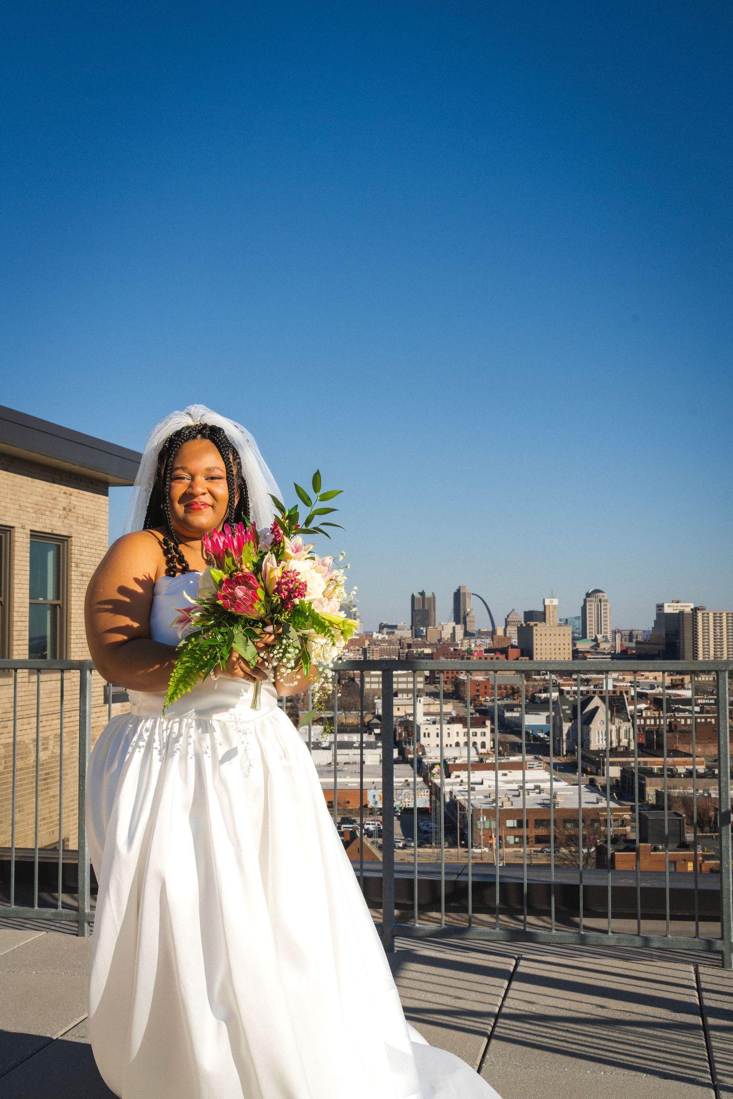 Radiant bride in white ball gown holding colorful bouquet on rooftop with city skyline and Gateway Arch in background.