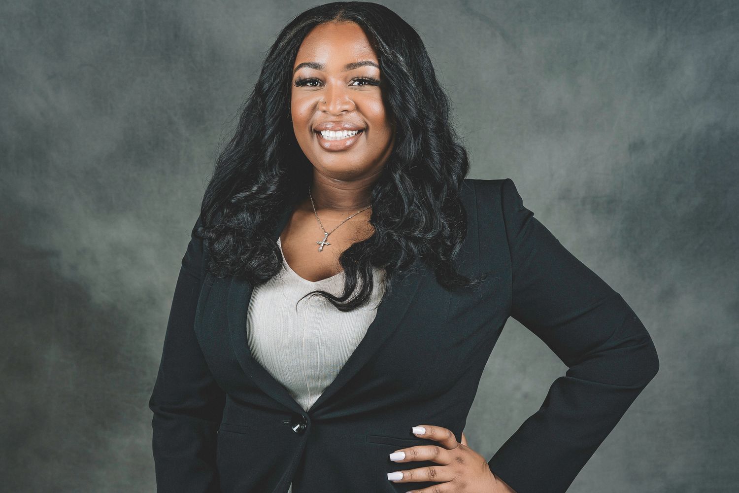 Confident woman in a black blazer smiles warmly while posing with hand on hip against a gray studio backdrop.