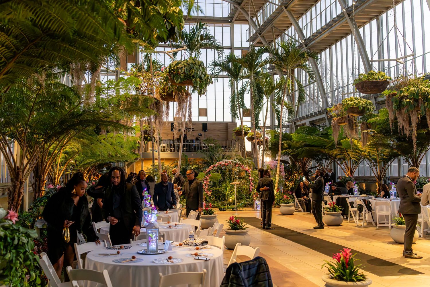 Elegant indoor garden event with lush tropical palms, hanging plants, and white-draped tables under a glass ceiling.