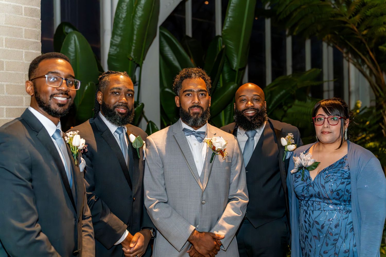 Groom in gray suit poses with groomsmen and bridesmaid in blue dress at wedding reception.