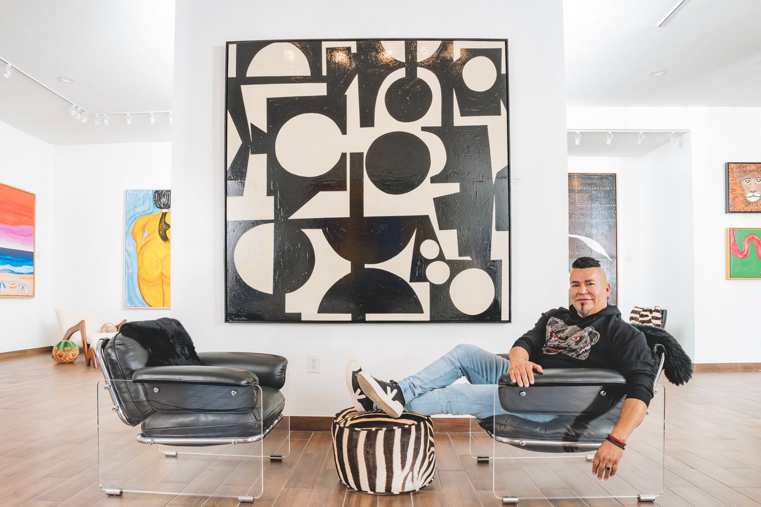 Artist relaxing in black leather chairs before large geometric black and white artwork in gallery.