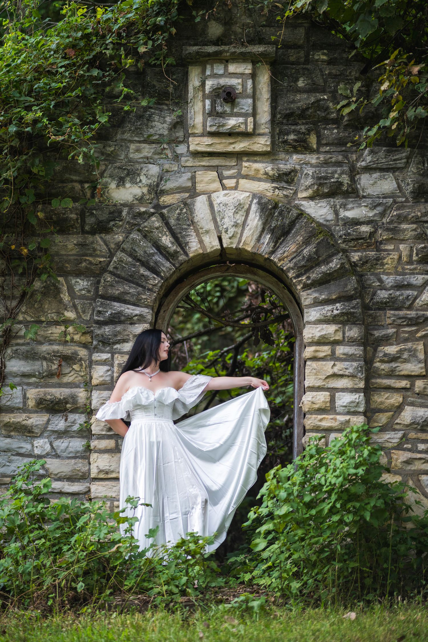 Woman in white off-shoulder gown posing in stone arch doorway surrounded by lush green ivy.