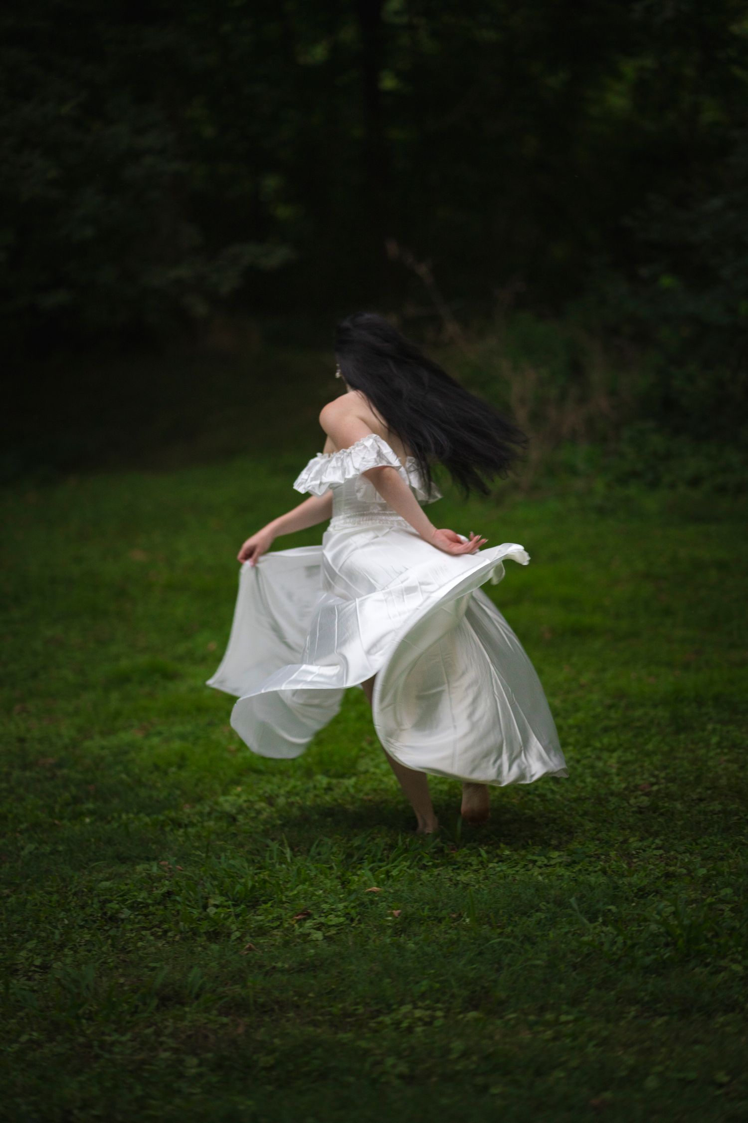 Woman in white off-shoulder dress twirls barefoot on green grass with flowing skirt.