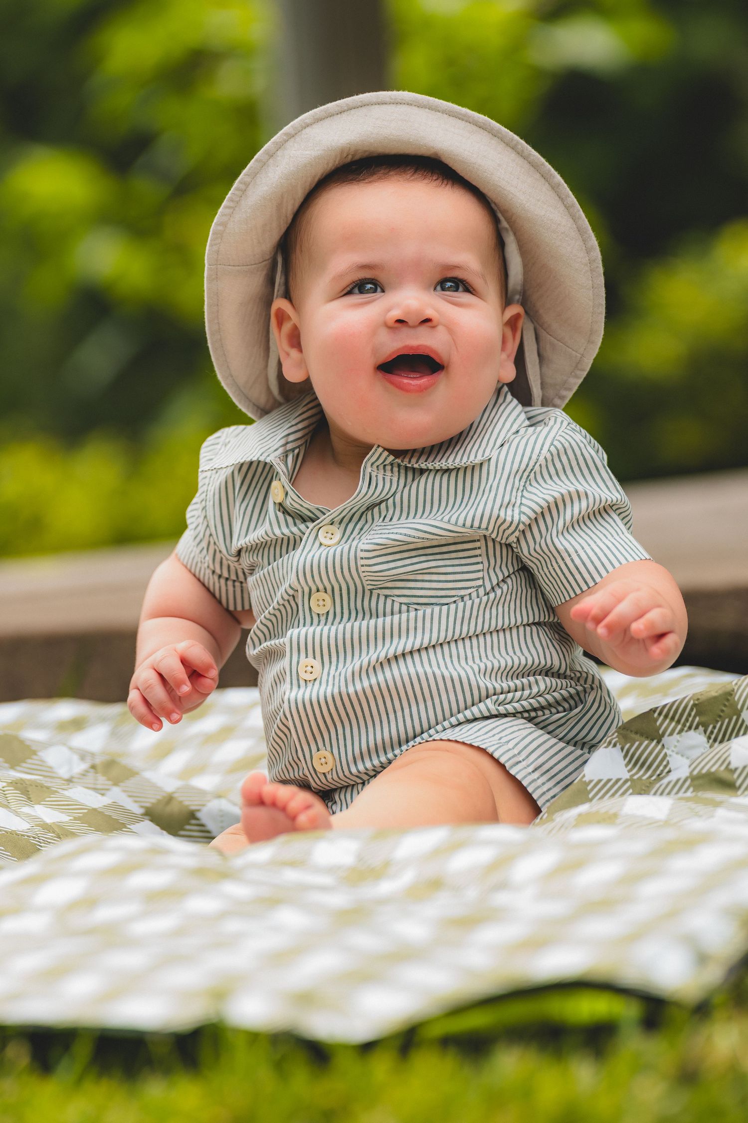 Adorable smiling baby wearing bucket hat and striped outfit sitting on checkered blanket outdoors.
