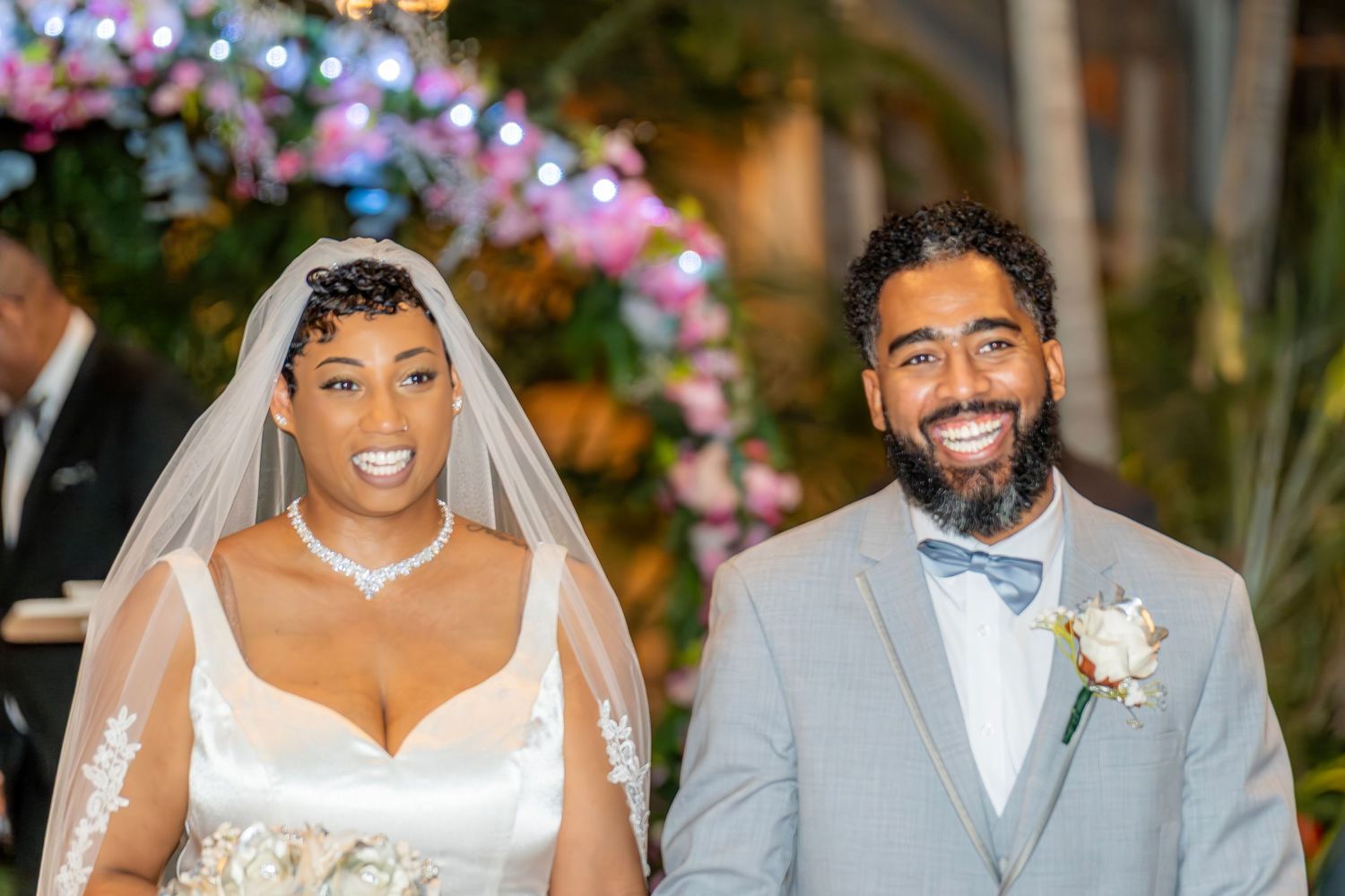 Newlyweds smile joyfully after ceremony, bride in white lace gown, groom in gray suit with boutonniere.