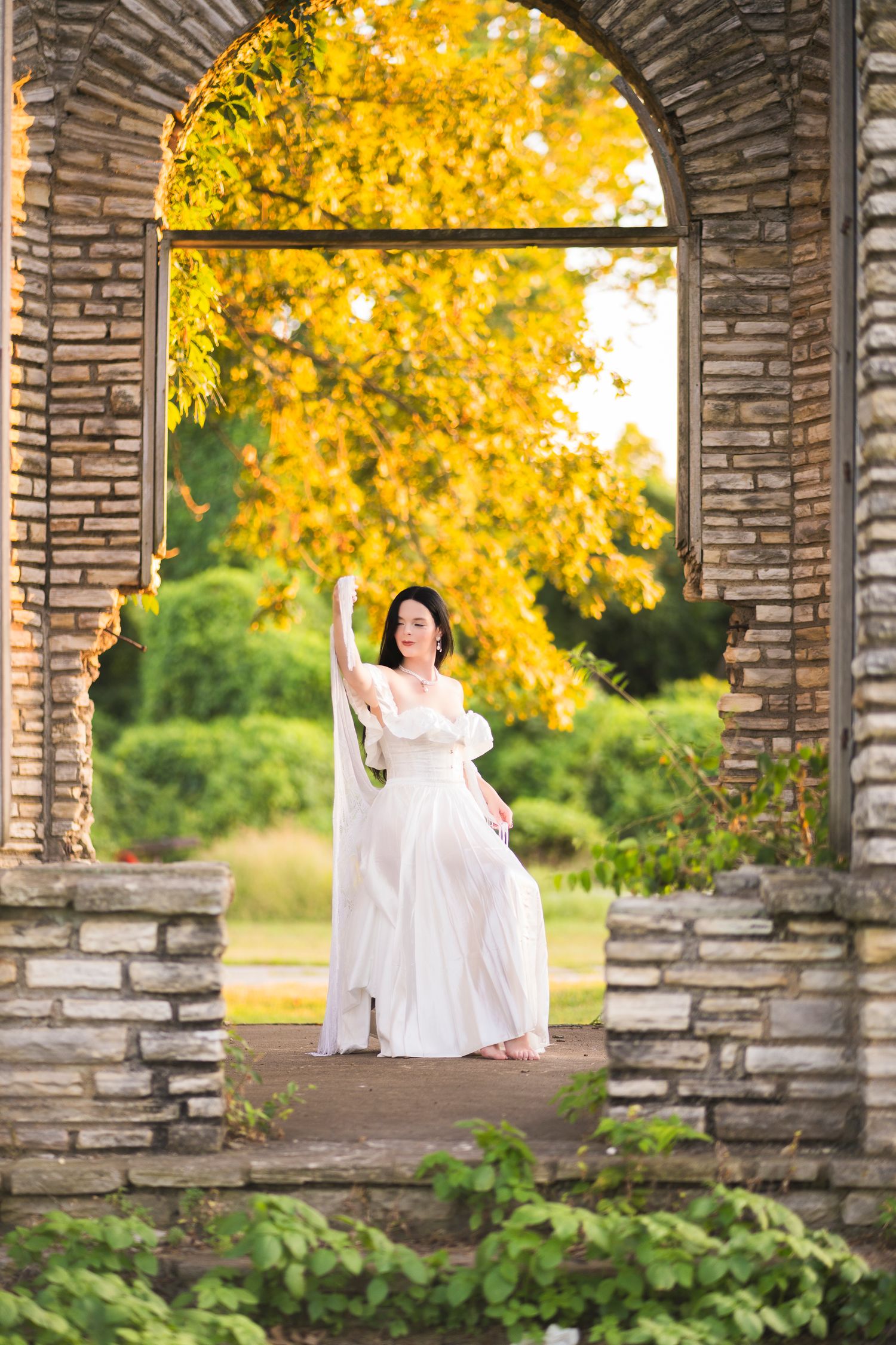Woman in sheer white maternity gown posing gracefully in brick archway with yellow autumn trees.