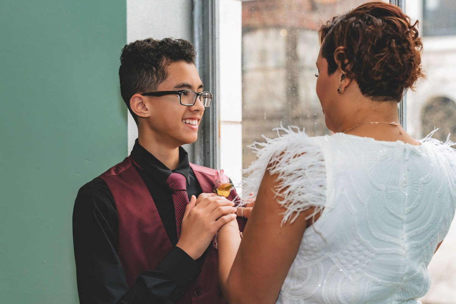 A smiling teenage boy in a burgundy vest gets his boutonniere pinned by a woman in a white dress.