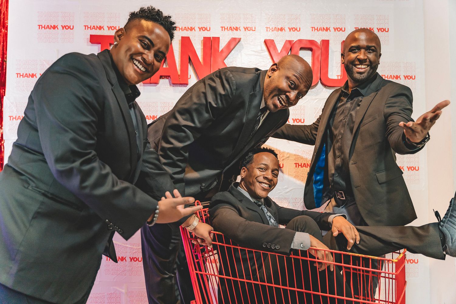 Four joyful men posing with shopping cart in front of Thank You event backdrop.