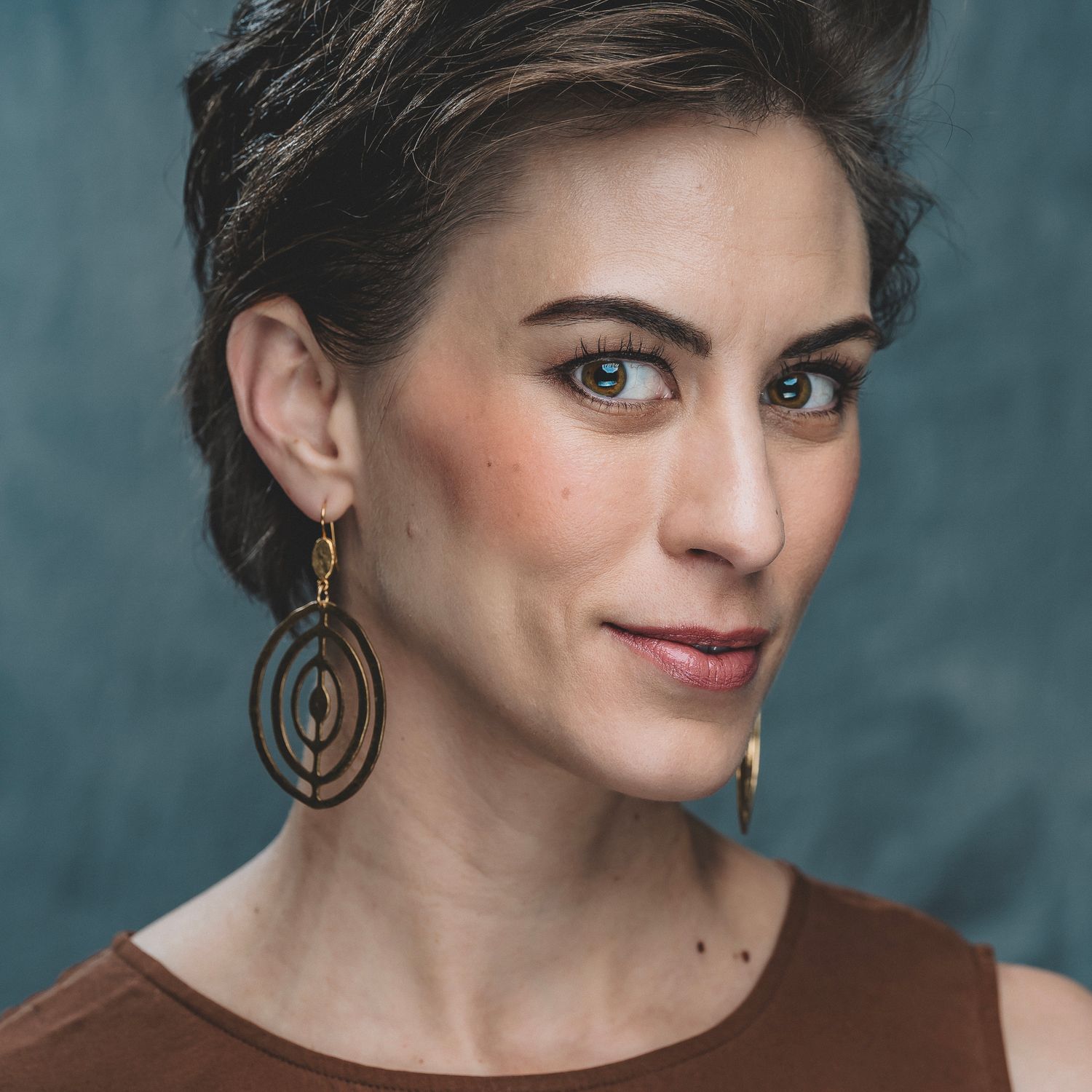 Dark-haired woman wearing layered geometric gold drop earrings in brown top.