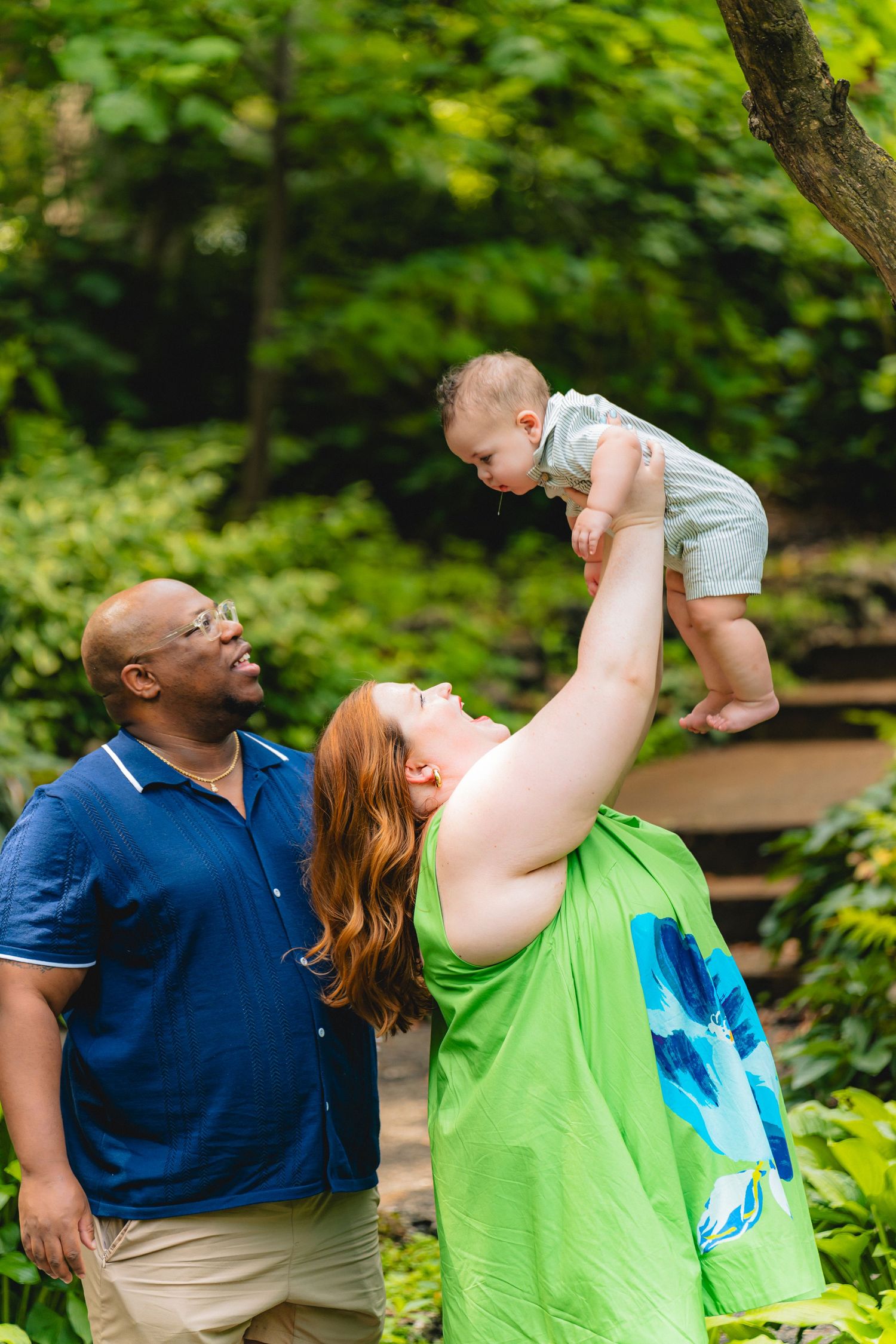 Interracial family outdoors, woman lifting baby overhead while man watches and smiles.