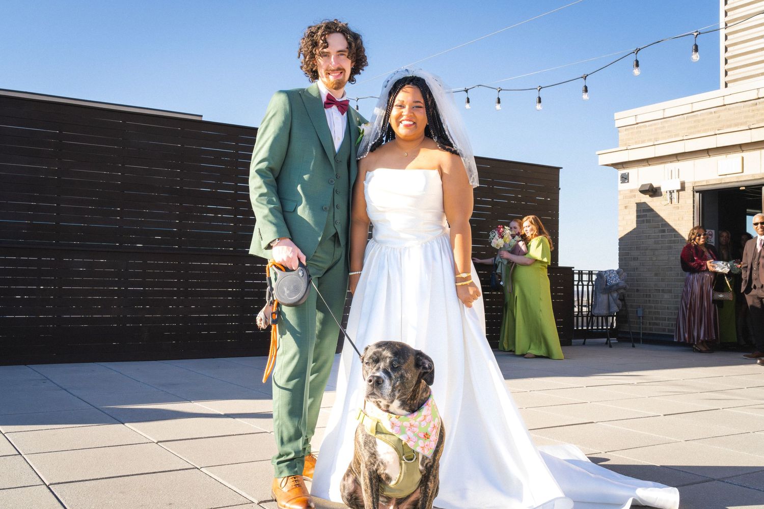 Newlywed couple posing with their bulldog on a rooftop terrace, groom in green suit and bride in white wedding gown.