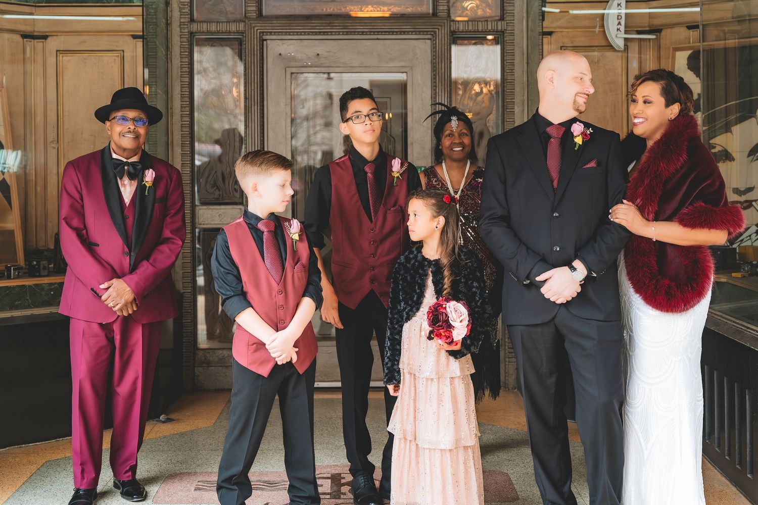 Wedding party stands together at venue entrance in coordinating burgundy and black formal wear.