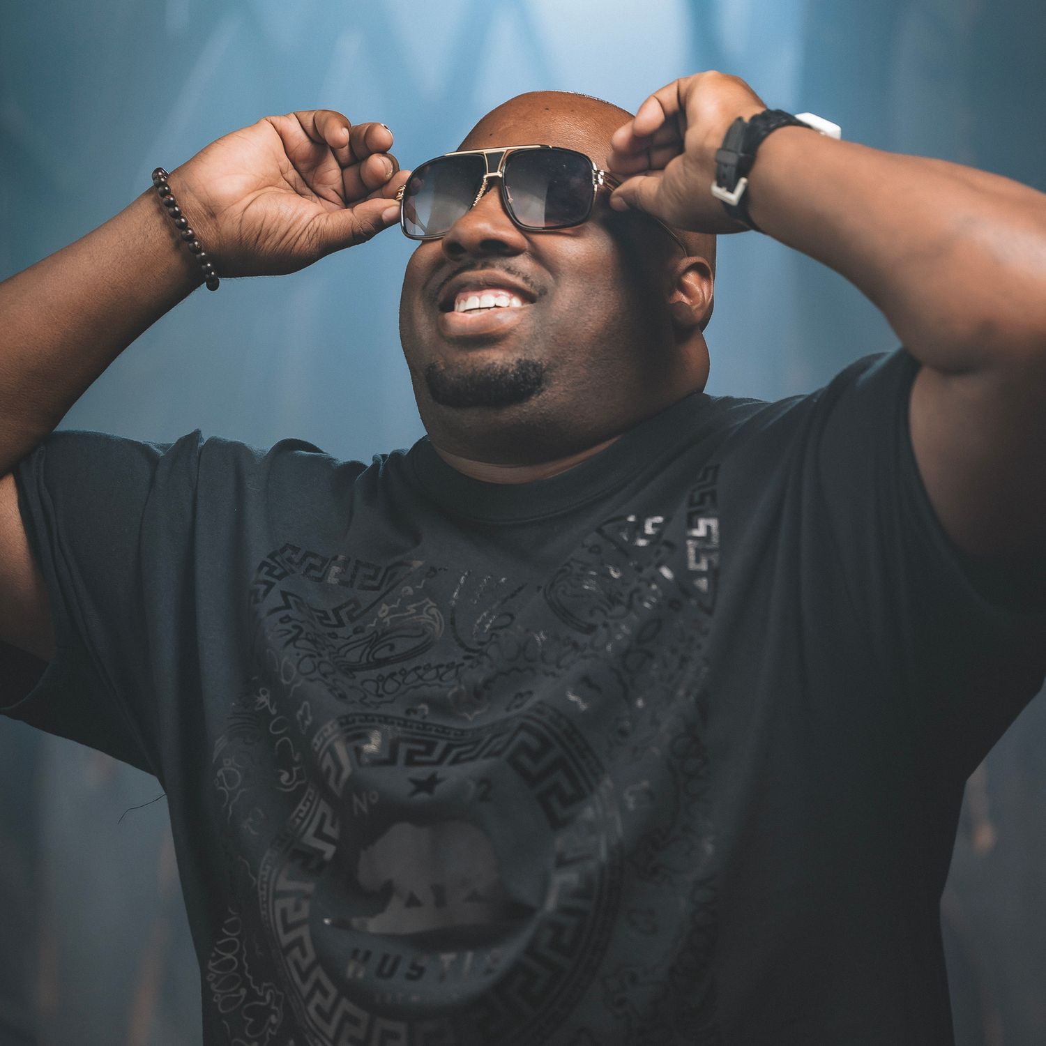 Performer in all black adjusts sunglasses on stage with dramatic blue lighting behind them.