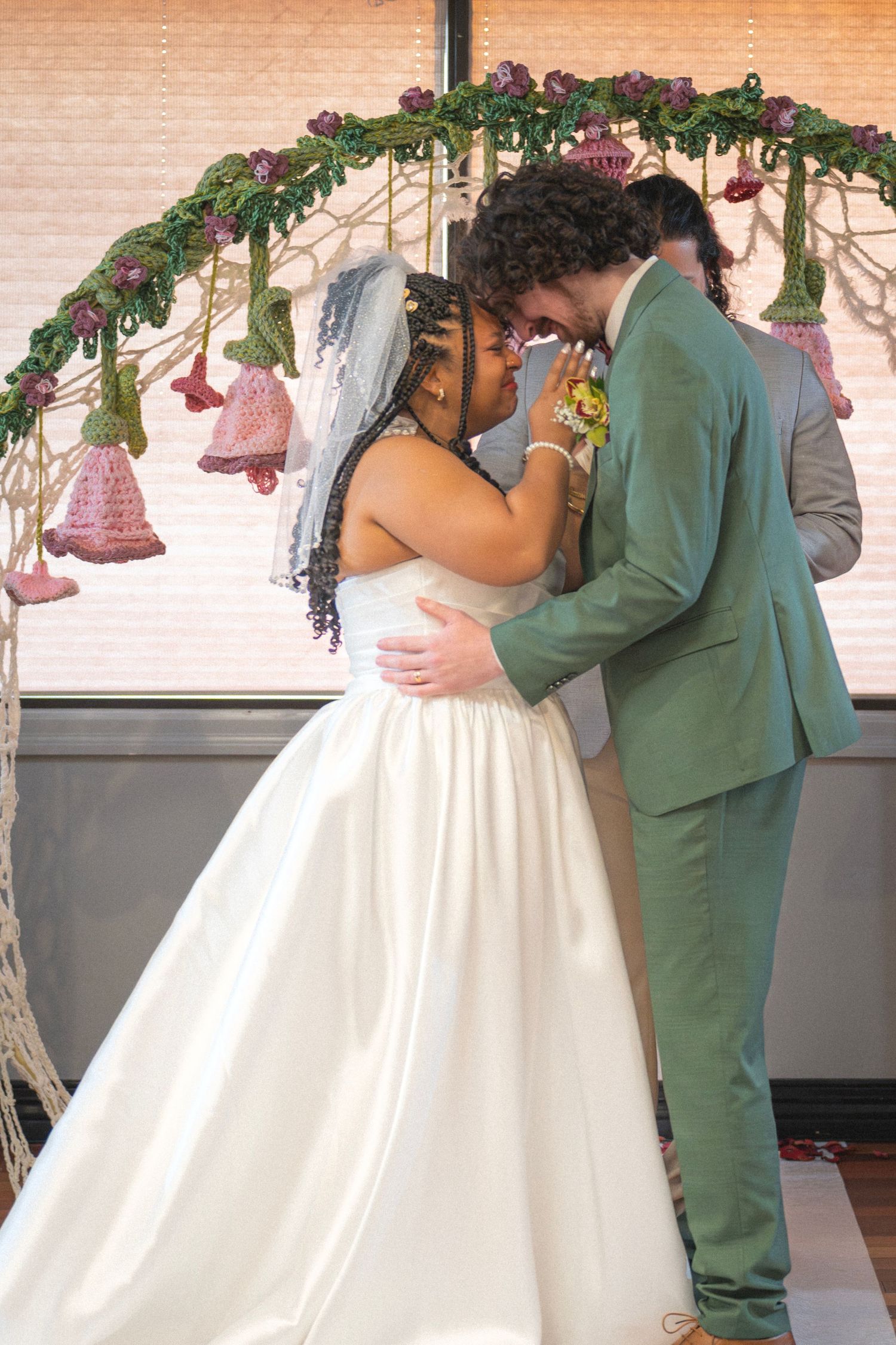 Bride and groom sharing an intimate first kiss moment under decorative floral arch, groom in sage green suit.