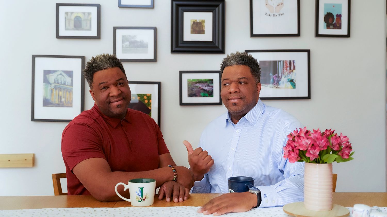 Two smiling men sitting together at a dining table with framed artwork on the wall behind them.