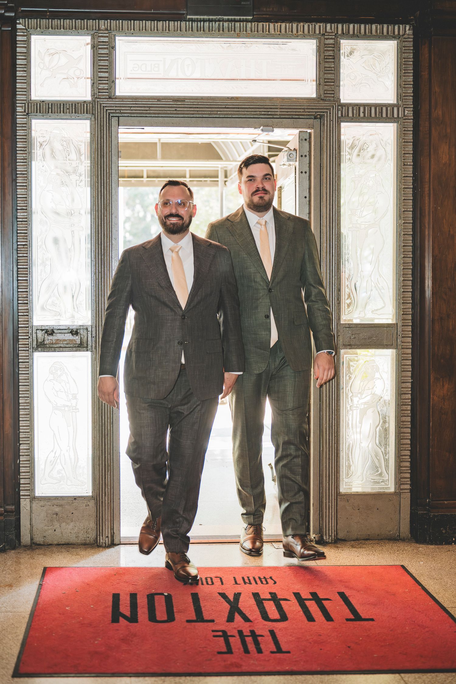 Two grooms in elegant gray suits walk through the grand entrance of The Thaxton Saint Louis.