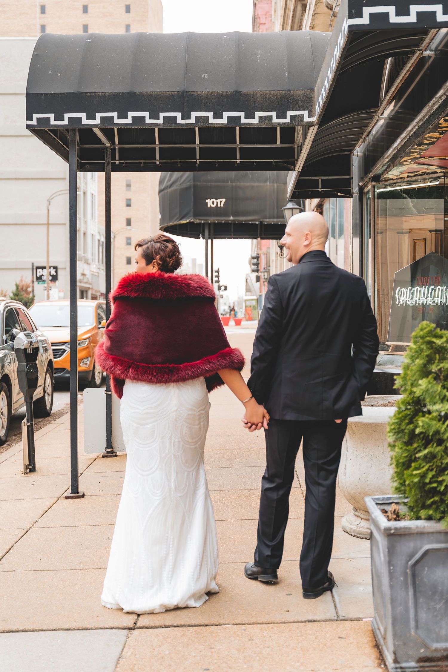 Bride and groom walk hand in hand down city sidewalk, bride wearing burgundy fur shawl.